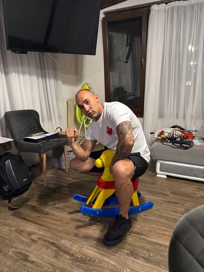 The image depicts an indoor scene in Hristo Danovo, Bulgaria. A light-skinned man with a shaved head and tattoos on both arms is centrally positioned, facing the viewer. He wears a white t-shirt with a red maple leaf logo on the left chest, black shorts, and black sneakers. He is seated on a child's yellow and red rocking horse with blue rockers, his right fist clenched and raised, while looking directly at the camera with a neutral expression.
To the man's left, a dark grey quilted chair holds a white and black object resembling a PlayStation 5 console and several wooden pencils. On the wooden laminate floor near the chair are a black hard-shell travel bag and two olive-green slippers. In the background, white sheer curtains cover a wall and a dark brown framed window or glass door. A large, black flat-screen television is mounted on the wall above the curtains on the left, with a small black digital receiver displaying "0135" beneath it, and white cables dangling. To the right background, a grey upholstered bench or sofa is visible, cluttered with various cables, a red box, and other small items. Behind the man, a green child's high chair or bouncer and a patterned fabric storage bin are present. The room has light-colored walls and a wooden ceiling beam.