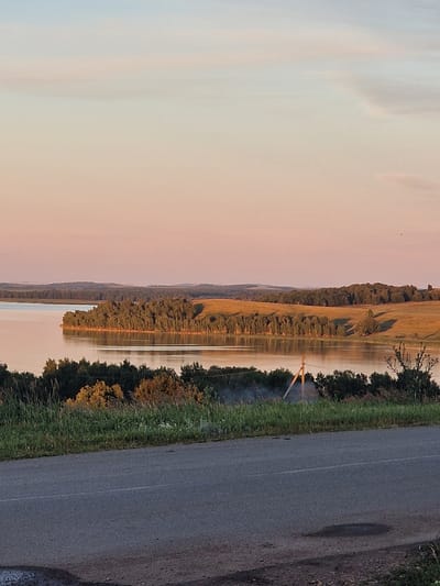 The image depicts a tranquil natural landscape at dusk. A calm body of water, likely a lake or river, occupies the left and central portions of the frame. The water reflects the soft hues of the sky, which transitions from a pale blue at the top to a warm pink and orange closer to the horizon. Along the far bank of the water, a dense line of trees forms a dark green border against the lighter landscape. Beyond the trees, rolling hills covered in golden-brown fields stretch towards the horizon, interspersed with more distant, darker forested areas. The foreground features a paved road, partially visible, with a grassy verge and scattered bushes along its edge. A simple wooden utility pole stands near the water's edge, with faint wisps of smoke rising from behind it. The overall atmosphere is serene and peaceful, with the soft light of sunset casting long shadows and bathing the scene in a warm glow. The image does not contain any elements that would suggest it is located in Kashgar City, China; rather, it appears to be a rural or natural setting in a temperate climate.