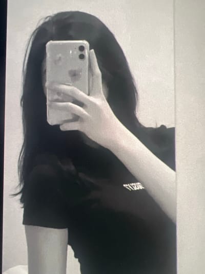 The image depicts a monochrome, chest-up mirror selfie of a person in an indoor setting. The individual holds a light-colored mobile phone with two visible rear camera lenses and a clear case, positioned to obscure their entire face. The person has long, dark hair, visible on both sides of the phone and extending over the shoulders. They are wearing a dark, short-sleeved t-shirt with light-colored, inverted text or a logo visible on the upper left chest area. The person's left arm and hand are visible, holding the phone. The background is a plain, light-colored surface, likely a wall or another mirror reflecting the scene. There are no visual elements within the image to identify the location as Ittenheim, France.