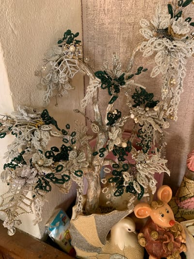 The image presents a close-up view of a beaded tree or decorative arrangement composed of white, silver, and green beads, some resembling leaves or flowers. The structure of the tree appears to be made of a silver-colored material. In the background, partially visible, are assorted objects including a ceramic mouse figurine, a bag of money, and a white porcelain duck. The objects rest on what appears to be a shelf with a textured surface. The setting appears to be an indoor space with muted lighting. The image was taken in a domestic environment. There is no information given in this image to connect it to the city of Slavyani, Bulgaria.
