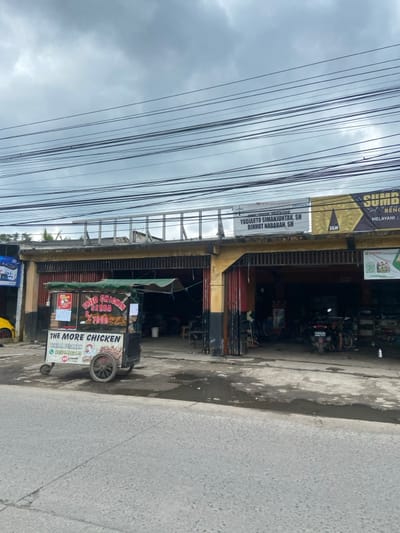 The image shows a street scene in Baleendah, Indonesia. There is a food cart parked on the left side of the street, with a sign that reads "Fried Chicken Rp. 7.000". The cart is in front of a yellow building with a red door. On the right side of the street, there is a large building with a sign in Indonesian, which appears to be an advertisement. Several power lines run across the scene. There are a few motorbikes parked in front of the building, and a person is walking towards the camera. The street is wet and has puddles of water. The sky is grey and cloudy.