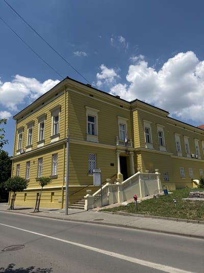 The image shows a yellow building with white trim and windows. The building is on a street with a sidewalk and a road. There are trees and grass in front of the building. The sky is blue with some clouds. The building appears to be a historical or public building. The flags on the balcony suggest it may be a government building. The building is located in Sremska Mitrovica, Serbia.