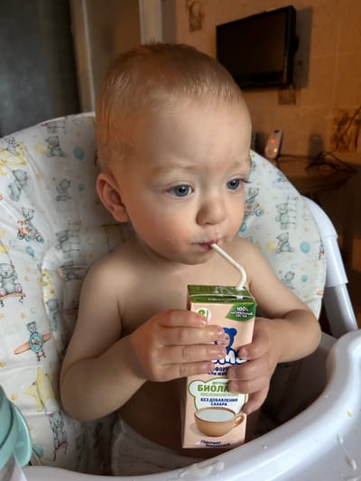 A toddler with light blonde hair and blue eyes is seated in a white high chair with a fabric liner patterned with teddy bears, airplanes, and balloons. The child, bare-chested and wearing a white diaper or underwear, is actively drinking from a small pinkish-beige carton through a white bent straw, with visible milk residue around the mouth. The carton, held by both hands, is labeled in Russian as "БИОЛАКТ" (Biolact), a "ДЕТСКИЙ КИСЛОМОЛОЧНЫЙ" (children's fermented milk) product "БЕЗ ДОБАВЛЕНИЯ САХАРА" (without added sugar) and with "100% НАТУРАЛЬНЫЙ СОСТАВ" (100% natural composition). The background reveals a light-colored tiled wall, a mounted black flat-screen television, a white device with a red indicator light, and black wires, indicating a domestic interior such as a kitchen or dining area in Voskresensk, Russia.