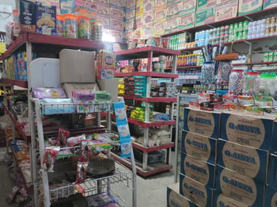 The image depicts the interior of a small grocery store in Meunasahbaet, Indonesia. The shelves are stocked with a variety of food items, including snacks, drinks, and household goods. Cardboard boxes of "Q-AISYA" brand mineral water are stacked on the floor. The shelves are crowded with products, and boxes of "Suksess" brand instant noodles are stacked high against the back wall.