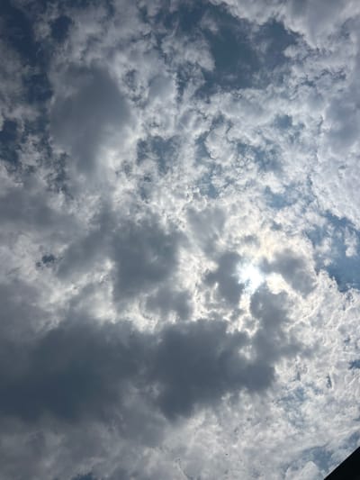 A low-angle, full-frame shot captures a cloudy sky above Giussano, Italy. The sun, partially obscured by dark, cumulus clouds, creates a bright, hazy glow in the upper right quadrant of the image. Scattered, wispy white clouds fill the rest of the frame, interspersed with patches of blue sky. The composition is entirely focused on the atmospheric conditions, with no discernible man-made structures or living beings visible. The overall impression is one of an overcast day with intermittent sunshine.