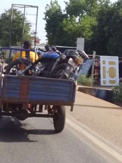 The image shows a blue flatbed truck driving on a paved road in Dungu, Ghana. The truck bed is heavily laden with what appears to be scrap metal, including several disassembled motorcycles. A man wearing a yellow and black striped shirt is in the bed of the truck, possibly securing the load. A white door with gold accents is resting against a pile of objects behind the truck. Trees and a framed structure are visible in the background. The scene appears to be within an urban environment.