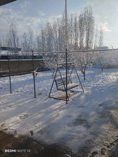 A snow-covered backyard features a metal swing set dusted with snow. Behind the swing, trees are also coated in snow, creating a winter scene. A brick wall runs along the back of the yard. The ground is covered in snow with footprints visible near the bottom of the frame. A clothesline stretches across the yard. The sky is partly cloudy with sunlight filtering through.