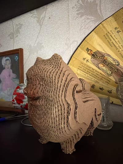 A cardboard sculpture of an animal, possibly a pig or bear, is positioned on a dark surface in front of a wall with floral wallpaper. To the left of the sculpture is a framed photograph of a baby wearing a pink outfit and a white headband. Behind the sculpture and to the right is a partially open, yellow fan with an illustration of a man in traditional clothing and Chinese calligraphy. A glass object, possibly a vase or cup, is visible to the right of the fan. The lighting suggests an indoor setting. The scene is consistent with a domestic environment in Shepsi, Russia.