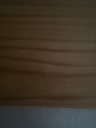 The image is a close-up, slightly blurred photograph of a light brown, horizontally-grained wooden surface. Subtle variations in color suggest the natural grain of the wood, with slightly darker lines running horizontally across the frame. The lower portion of the image shows a lighter, textured surface that appears to be fabric or a similarly textured material, possibly a wall or upholstery. There are no people, objects, or discernible elements that would indicate a specific location within the city of Нарынка, Russia.