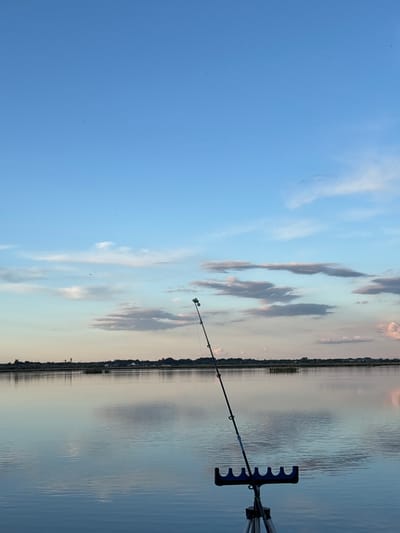 The image depicts a fishing scene at a body of water, likely a lake or wide river, in Khryashchevka, Russia. A fishing rod is visible extending towards the water, supported by a rod rest. The sky is partially cloudy. On the opposite shore, vegetation and buildings are discernible. The water surface shows subtle ripples.
