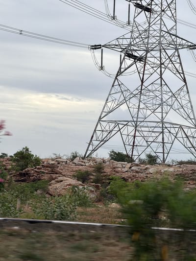 The image depicts an outdoor scene dominated by a large, industrial-style electricity pylon. The pylon is constructed from intricate metal lattice work and supports multiple high-voltage power lines that stretch across the frame. The pylon is situated in a rocky, arid landscape with scattered vegetation, including bushes and some trees. In the foreground, a blurry element suggests movement, possibly from a vehicle. The sky is overcast with a grey, cloudy texture. The scene is likely located in or near Nannur, India, given the context provided, and is consistent with rural or semi-industrial areas found in such locations.