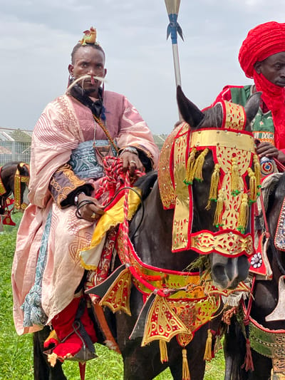 The image depicts two men on horseback, likely captured in Kafanchan A, Nigeria, given the prompt. Both horses are adorned with elaborate, traditional ornamentation, including gilded and colorful fabrics. The rider closest to the viewer is wearing a light pink and patterned robe and possesses distinctive facial decorations, including white elements near the nose and ornate headwear. The second rider, behind and slightly above, is wearing a red turban. One rider holds the reins of the horse and displays jewelry on his fingers. The background shows green vegetation and a cloudy sky.