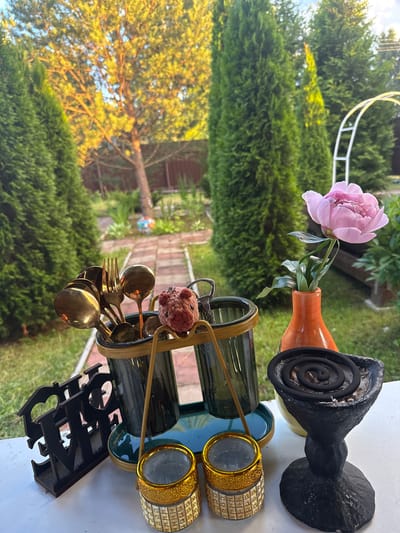 This media file depicts an outdoor still life, likely a table setting in a garden. The scene is oriented vertically, with the top of the image appearing on the left. In the foreground, a set of golden cutlery, including spoons and a fork, rests in a metallic holder. A small crocheted pig charm is attached to the holder. Behind the cutlery, two dark green glasses are visible, placed on a dark green tray with a gold rim. To the right of the glasses are two decorative items that resemble candle holders or napkin rings, featuring intricate gold and diamond-like patterns.
Further back, a bright pink peony in a small orange vase adds a touch of color to the arrangement. Next to the vase, a black, coiled object, possibly an insect repellent coil, sits in a decorative black holder. In the background, lush green trees and foliage create a natural setting. Sunlight illuminates the scene, suggesting it's daytime, possibly late afternoon given the warm lighting. The ground appears to be paved with bricks, and a white, possibly metal, garden arch is partially visible. There is no visible text in the image. The overall impression is one of a prepared, decorative outdoor space, possibly for an event or a tranquil moment. The location context provided is Тмз Снт, Russia.