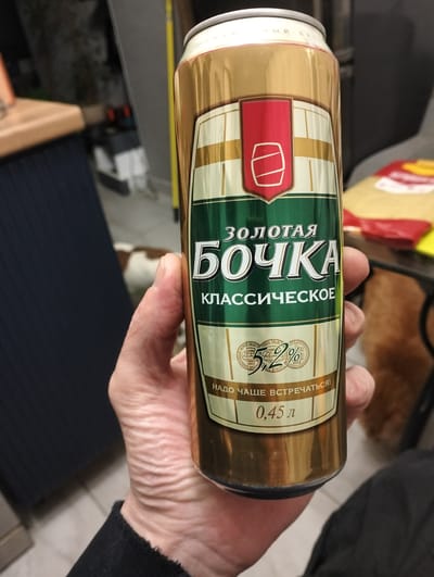 A person is holding a golden beer can. The can has green and white labeling with Russian text and a red emblem featuring a barrel. The text on the can includes "ЗОЛОТАЯ БОЧКА" (Golden Barrel), "КЛАССИЧЕСКОЕ" (Classic), "5,2%", and "0,45 л" (0.45 L). The background is blurred, showing what appears to be an indoor setting with furniture and possibly other objects. The location is stated to be in the city of Zyuzino, Russia.