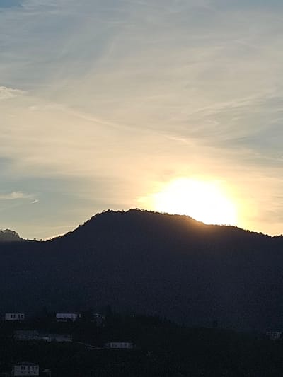 The image presents a serene landscape scene captured at either sunrise or sunset, with the horizon line running vertically across the middle of the frame due to the image's orientation.
The sky, occupying the left half of the image, displays a beautiful gradient of colors, transitioning from light blue at the top to vibrant warm yellows and oranges near the sun. Wispy, ethereal clouds are scattered across the sky, illuminated by the intense light. The sun itself is partially visible, a brilliant, glowing orb peeking just above the crest of a large mountain, casting a strong backlight.
The right half of the image is dominated by the dark silhouette of a vast mountain or large hill. Its rugged, irregular outline is sharply defined against the bright sky, emphasizing its imposing presence. The mountain appears heavily forested or covered in dense vegetation, contributing to its dark, shadowed appearance.
In the lower right corner, at the base of the mountain, several small, light-colored buildings are visible. These structures are somewhat indistinct but indicate human habitation within the vast natural setting.
The scene evokes a sense of peace and natural grandeur. The time of day is clearly either dawn or dusk, characterized by the low sun and warm, glowing light. The weather appears clear with delicate cloud formations. There are no people visible, and no specific activities are taking place. No visible text is present in the image. The location context provided is ქვედა სამება, Georgia.
