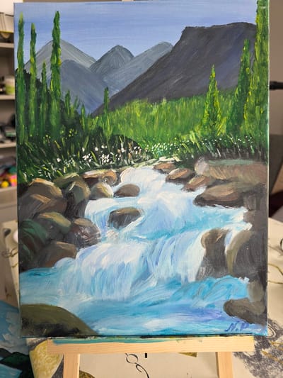 A painting depicts a natural landscape with a focus on a flowing river. The scene includes mountains in the background, lush green grass and trees on the banks, and a body of water with rocks. The water is rendered with white and blue brushstrokes to convey movement and foam. The foreground shows rounded rocks of various earthy tones. The sky is a clear blue. The painting is on canvas and is displayed on a wooden easel. The bottom right corner of the canvas shows a signature that appears to be "N.P."
This image does not depict any identifiable location in the city of Claremorris, Ireland.