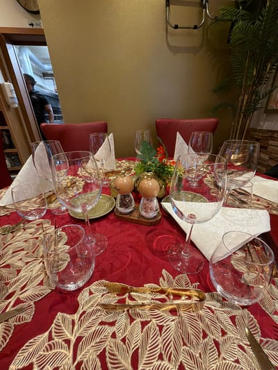 The image depicts a formally set dining table in Fentange, Luxembourg. The table is covered with a red tablecloth featuring an intricate gold leaf pattern. Six wine glasses and six water glasses are arranged at settings with folded white napkins. A centerpiece with floral arrangements, salt and pepper shakers rests in the middle of the table. Red upholstered dining chairs surround the table. In the background, a partially visible individual leans into a maintenance access area. A "WC" sign and pipes are visible in the background.