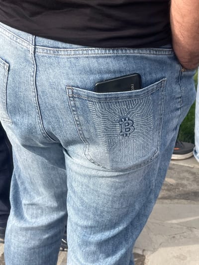 A close-up shot focuses on the lower torso of a person wearing light blue denim jeans. A smartphone, with the partial word "red" visible, is partially inserted into the rear right pocket of the jeans. The pocket features an embroidered design that incorporates a "B" and what looks like circuit lines. The individual is wearing a black garment tucked into the jeans. The background consists of a textured surface, possibly a sidewalk, with additional details including parts of footwear and what may be a concrete structure visible further in the frame. The location is likely outdoors during daylight hours. The city and country, Akhuryan, Armenia, cannot be independently confirmed.