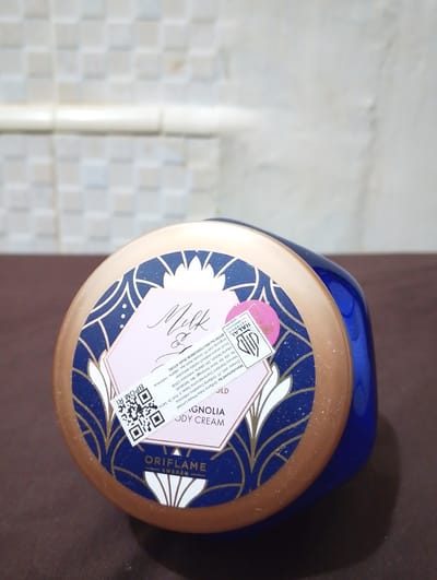 A close-up, slightly angled shot shows the lid of a blue Oriflame body cream jar with a gold rim. The lid features a decorative blue and gold pattern. A pink label on the lid has text and a QR code. The text on the label appears to include "Milk & Magnolia Body Cream," "HALAL," and manufacturing information that mentions "Manufactured by: Oriflame India Private Limited." Also visible is "BPOM RI" next to the QR code, which is a regulatory agency in Indonesia. The background is a textured, off-white wall and a brown surface. No people are present. The scene is indoors, possibly a bathroom or dressing area. The location in Geulanggangbaro, Indonesia is not depicted in the image.