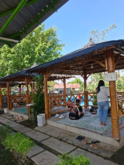 The image shows a scene in Kecamatan Tulung, Indonesia, located at latitude -7.592 and longitude 110.65. It features a bamboo hut structure with a thatched roof. The hut is open on all sides, providing views of a swimming pool and lush greenery. There are three people present: a woman in a light blue shirt and blue jeans standing near the hut, and two people sitting on a mat inside the hut. The individuals sitting on the mat are wearing casual attire, and one of them is obscured by a black and white patterned bag. The hut is situated on a paved pathway bordered by green plants and trees, providing shade from the sun. The scene conveys a sense of leisure and relaxation, with the people seemingly enjoying their time in the outdoor space. The image appears to be taken during the day, under a bright blue sky.