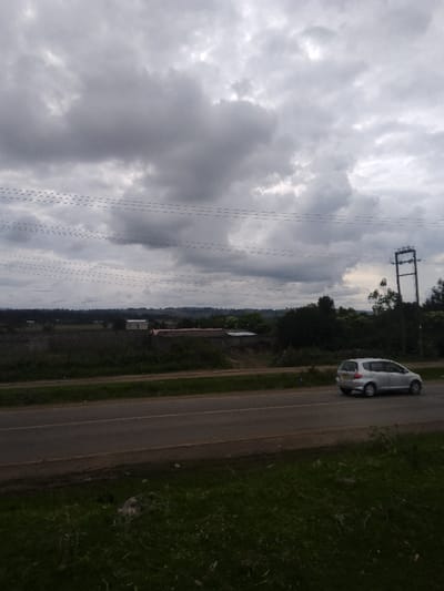 The scene depicts an outdoor landscape in Njoro ward, Kenya, under an overcast sky dominated by heavy grey clouds. A two-lane asphalt road traverses the mid-ground horizontally. A silver hatchback vehicle, consistent with a Honda Fit, is observed on the right lane, traveling from right to left. The immediate foreground features a dark green grassy verge. Beyond the road, to the left, are open fields extending into the distance. In the central background, several low-rise structures are visible; one possesses light-colored walls and roof, while others exhibit darker, earthy construction. To the right, a cluster of dense trees and a tall, dark metal electricity transmission tower are present. Multiple parallel overhead power lines are strung across the upper portion of the frame, connecting to the transmission tower. Distant elevated terrain is visible on the horizon beneath the clouds. No people are visible.