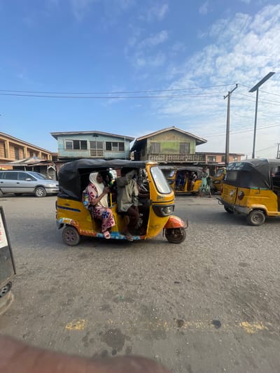 In Akowonjo, Nigeria, a sunny daytime scene captures a busy street with several yellow auto-rickshaws, also known as "keke napep." In the foreground, a yellow keke napep is prominently displayed, carrying two passengers. A woman in a vibrant patterned dress and headscarf sits in the passenger seat, while a man in a light-colored shirt and dark trousers drives. Behind them, another yellow keke napep is visible with passengers. Further in the background, more auto-rickshaws are present, suggesting a bustling mode of transportation. The street is paved, and buildings with varying architectural styles line the sides, including a two-story structure painted blue and another with green accents. Power lines and streetlights are also visible, indicating the presence of urban infrastructure. The sky is blue with scattered white clouds.