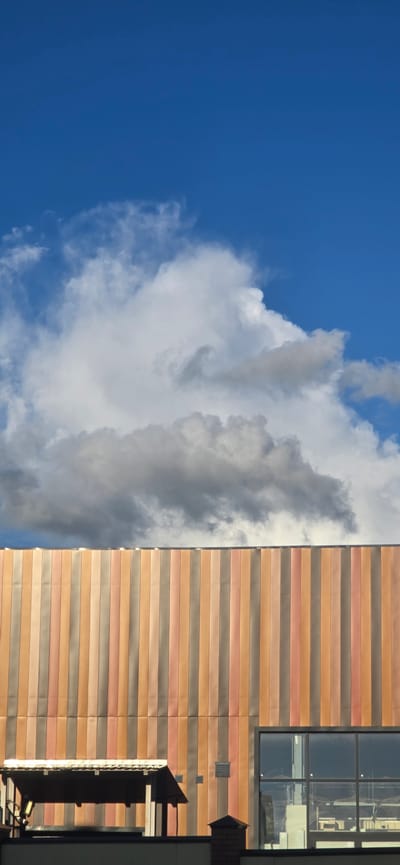 The image shows a building with a facade made of vertical panels in various shades of brown, orange, and pink, set against a bright blue sky with white and gray clouds. A portion of a covered outdoor area with a slanted roof is visible in the lower left, and a large window with reflections is in the lower right. The scene is set in the village of Russkaya Derevnya-2 KP, Russia.