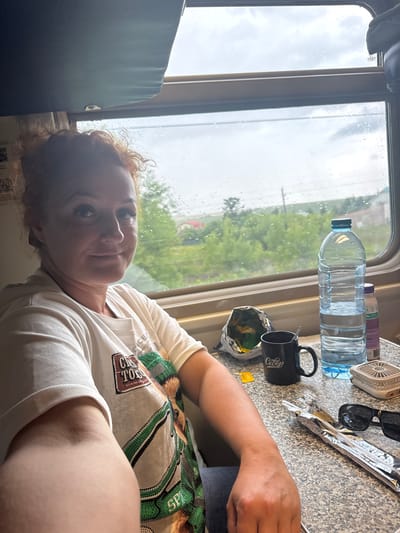 An eye-level, medium shot captures a woman with curly red hair pulled into a bun, looking directly at the camera. She is seated at a table inside a train car, with a window to her right revealing a landscape of green trees and buildings under a cloudy sky. The woman is wearing a white t-shirt with a graphic print featuring a dog and green accents. On the table in front of her are various items: a large plastic bottle of water, a black mug with Cyrillic lettering, a crinkled bag of snacks, a yellow tea bag, a pair of black sunglasses, a folded silver foil wrapper, and a small white fan. The table has a speckled gray and black surface. The train car interior is visible with a dark blue sunshade above the window.