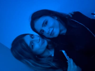 Two young women pose close together under a blue light. The woman on the left holds a phone. The other woman wears a dark top with a silver chain visible. Both have long hair and are gazing toward the viewer. The image is slightly angled, and the blue light creates a strong color cast.