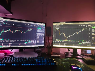 The image depicts a desk in a home office in Kfar Yona, Israel. Two computer monitors are displaying stock market charts. One monitor is on the left and one is on the right, with a keyboard and mouse in front. There is a pink fabric on the left side of the frame. There is a keyboard and mouse in the foreground. The background is a dimly lit room with pink walls. The desk is organized and clean. The monitors are displaying the charts of the day, with one showing a clear upward trend. The room is lit with a pink light coming from the walls, while a blue light is coming from the computer monitors.