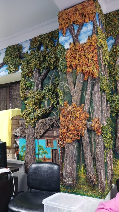 A bas-relief mural depicting an autumn forest scene with a log cabin is mounted on a wall and extends around a corner. The mural features textured trees with green and orange foliage, a rustic log cabin with blue walls, and a painted sky with clouds. A black office chair is positioned in front of the mural, partially obscuring the lower section. To the left of the chair, a desk with a computer monitor, a white power strip, and a clear plastic container are visible. A stack of brown cardboard boxes and a yellow piece of fabric are also present in the background. The setting appears to be an indoor space, possibly an office or workshop, in the city of Izberbash, Russia. There are no people clearly visible in the frame.