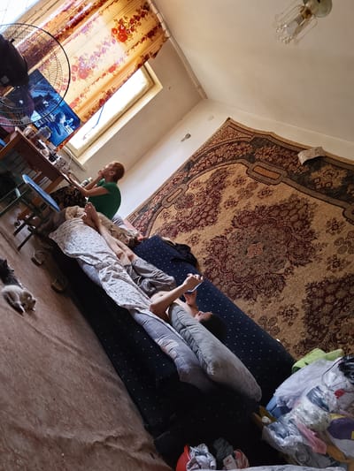 In a dimly lit room with a warm, patterned rug covering most of the floor and a large portion of the wall, two people are seen relaxing. One individual, with short red hair, is seated at a wooden table near a window, looking at a laptop or tablet. The other person is lying down on a sofa, also looking at a phone. The room is cluttered with various items, including a fan, a lamp on the ceiling, and a pile of clothes in plastic bags. Two small kittens are visible on the floor near the sofa, one of them lying down. The scene appears to be indoors, possibly a home, with natural light coming from the window. The overall atmosphere suggests a casual and relaxed setting. The image does not contain any visible text.