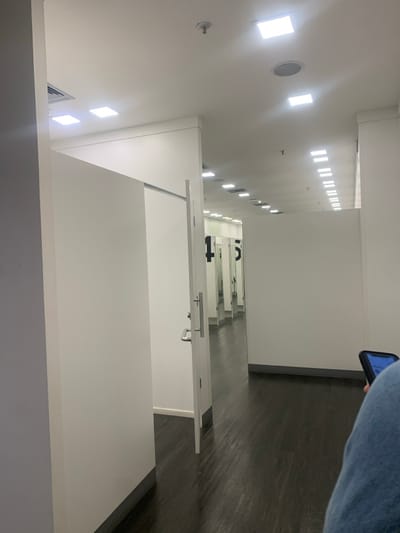 The scene is a brightly illuminated retail changing room area, located in Lauro de Freitas, Brazil. A long corridor is present, featuring a dark brown laminate floor and white walls. The white ceiling contains numerous recessed square LED lights, some circular fixtures, and a smoke detector. On both sides of the corridor, individual changing rooms are evident, each with white doors equipped with silver handles. Several doors are slightly ajar, revealing internal mirrors reflecting overhead lighting and the numbers "1" and "5," denoting changing room identification. A partial view of an individual is observed in the foreground on the right, clad in blue denim and holding a dark smartphone displaying an illuminated blue screen.