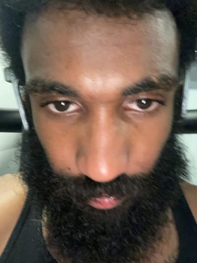 A close-up, slightly blurred, rotated portrait of a dark-skinned person with a thick, dark beard and dark eyes. The person has a shaved head. The background is indistinct, appearing to show a wall. The location in Aubervilliers, France cannot be determined from the provided image.