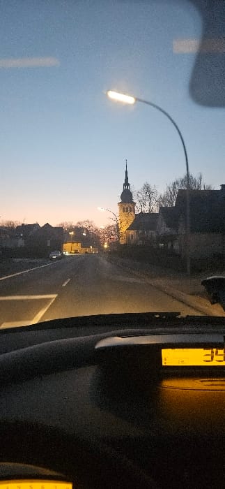 The image displays an exterior street scene in Rietberg, Germany, captured at dusk or dawn from within a vehicle. The sky transitions from light blue overhead to a subtle orange and pink hue near the horizon. In the immediate foreground, the vehicle's dark dashboard is visible, along with a portion of a yellow-illuminated digital display on the right showing the number "35." Reflections are apparent on the windshield. A modern LED streetlight on a curved pole is illuminated in the upper center, casting light.

A paved road, with a solid white line delineating its left edge, recedes into the distance. Along the right side of the road, the prominent, artificially illuminated spire and facade of St. Laurentius Church are visible, framed by bare trees. Various buildings, consistent with a town environment, line both sides of the road. A single vehicle with illuminated headlights is observed on the left side of the road further down the street. No individuals are discernible within the frame.