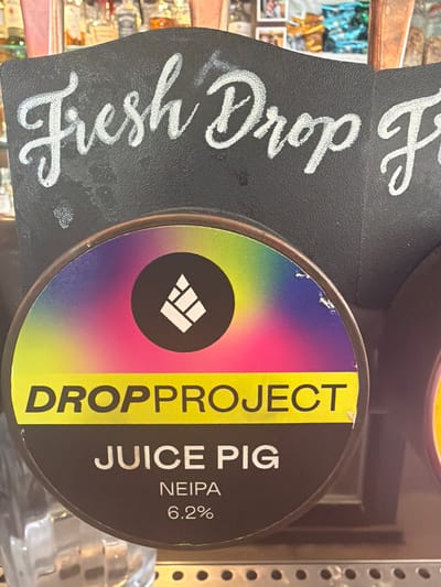 The image shows a close-up of a beer tap at a bar, likely in Epsom and Ewell, United Kingdom. The tap has a circular label with a colorful gradient and a white logo. Below the logo, the words "DROPPPROJECT" are written in yellow text on a yellow band. Further down, the text "JUICE PIG" is written in white. Below that, "NEIPA 6.2%" indicates the type of beer and its alcohol content. The background shows a chalkboard with white handwritten text that appears to say "Fresh Drop P". Behind the tap, blurred bottles of alcohol and bar equipment are visible.