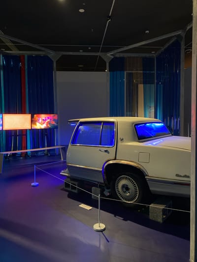 A white Cadillac sedan, seemingly an older model, is displayed inside a museum or exhibition space. The car is positioned on top of dark grey concrete blocks. The interior of the car, particularly the windows, is illuminated with a vibrant blue light. In the background, there are large screens displaying what appear to be video content. The screens are placed in front of blue and red vertically striped curtains, suggesting a deliberate artistic or thematic presentation. The environment is dimly lit, with spotlights highlighting the car and screens. The floor is a dark, reflective surface. The overall impression is one of a curated exhibit, possibly exploring themes related to automotive culture, history, or perhaps cinematic representations involving cars. The car's model name, "Sedan de Ville," is visible on the rear quarter panel. No people are present in the immediate vicinity of the car or screens.
