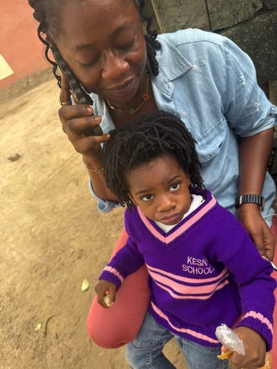In Ganyingbo, Nigeria, a Black woman is on a phone call, holding a dark-colored smartphone to her left ear with her right hand. She is wearing a light blue denim shirt with distressed details and a gold necklace with clover pendants. To her right, a young Black child sits on her lap, wearing a purple V-neck sweater with pink stripes and the text "KESN SCHOOL" embroidered on the left chest. The child is looking directly at the camera with a serious expression and holds a piece of bread wrapped in plastic in their right hand. The child is also wearing blue jeans. The background consists of a textured, sandy ground and a muted orange wall. The scene is captured from a high-angle perspective, looking down on the subjects.