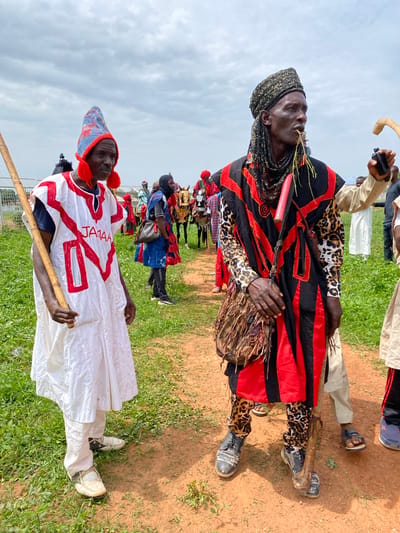 In Kafanchan A, Nigeria, two men stand out in a crowd on a field of green grass bordered by a patch of red dirt. The sky is overcast.
One man wears a predominantly white robe decorated with red patterns and the word "JAMAA" written on it. He holds a wooden staff. The other man wears a black and red patterned shawl over a leopard-print top and pants. He holds a bundle of twigs near his mouth and carries a mask.
A crowd of similarly dressed individuals can be seen in the background.