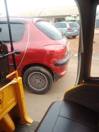 The image shows the interior of a yellow auto-rickshaw. The rickshaw is parked on a street in Abuja, Nigeria, specifically at the coordinates 8.955 latitude, 7.552 longitude. The rickshaw is facing a red Peugeot 206, parked on the street. The 206 has a damaged rear bumper and a silver-colored wheel cover. The rickshaw is empty, while the car is also empty. The background shows a gray and white building with a flat roof.  The sky is overcast, and there are buildings in the distance.  The environment is urban, with buildings lining the street.