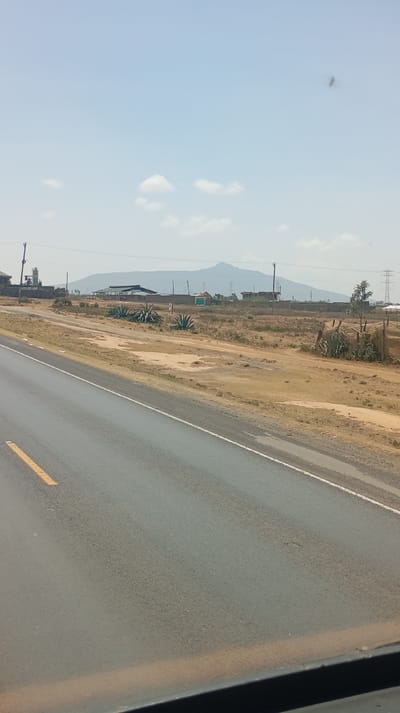 This image depicts a wide-angle view of a semi-arid landscape under a clear, light blue sky with scattered white cumulus clouds, consistent with the Hells Gate ward in Kenya.
In the foreground, a dark asphalt road occupies the lower left to mid-right of the frame, showing a solid yellow line on its left edge and a broken white line further right. The very bottom of the image reveals a dark, flat surface, likely a vehicle's dashboard or window frame.
The midground features a vast expanse of dry, brownish-yellow ground with patches of bare earth. Several large, spiky, succulent plants, resembling Agave, are sparsely distributed across this terrain. Towards the left, a concrete industrial structure is visible with what appears to be a silo or processing unit. Further right, a cluster of low-rise buildings with light-colored walls and dark, possibly corrugated, roofs are present, along with smaller, simpler structures, one featuring a vibrant green wall. On the far right, a wooden framework partially covered with a light-colored mesh is observed. Utility poles carrying power lines traverse the scene at various points.
In the background, a large, undulating mountain range or prominent hills are silhouetted against the horizon. High-voltage power transmission towers are visible towards the right base of these distant hills. No people are visible within the frame, and no specific actions or interactions are depicted. The overall impression is an open, undeveloped to semi-developed area with elements of utility infrastructure.