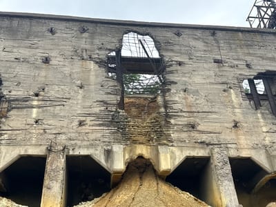 The image depicts a section of a severely damaged concrete structure, likely an industrial building or processing facility. The rough, gray concrete walls are heavily weathered and show signs of significant deterioration, with exposed rebar and large holes.

**Subjects:** The primary subject is the ruined concrete structure itself, characterized by its rough texture, broken rebar, and large openings. There are no visible people or animals.

**Objects:** The most prominent objects are the concrete walls, metal rebar, and square metal plates embedded in the concrete near the top. In the background, a glimpse of industrial metal scaffolding is visible against the sky.

**Scene/Setting:** The scene is one of destruction and decay. The setting appears to be an outdoor industrial area. The large openings in the wall reveal glimpses of the sky and some greenery, suggesting trees or other vegetation outside. The lower part of the image shows a large pile of light brown granular material, possibly earth, sand, or debris, accumulated in front of arched openings at the base of the structure.

**Activity/Event:** The image captures a static scene of a structure that has undergone significant damage, likely from an explosion, collapse, or prolonged neglect. There is no active event taking place in the frame.

**Notable Details:**
*   **Time of Day/Weather:** The sky is overcast, indicating a cloudy day with diffused lighting.
*   **Location Cues:** While not explicitly visible in the image itself, the provided location context is Kapan, Armenia, suggesting this is likely a historical or industrial site within that region.
*   **Emotions:** The image evokes feelings of decay, ruin, and perhaps a sense of past industrial activity now lost.

**Visible Text:** There is no visible text in the image.
