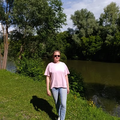 A woman wearing a pink t-shirt and blue jeans stands in a grassy area next to a body of water. She has sunglasses on and her hair is pulled back. The scene is outdoors with lush green trees and foliage surrounding the water. The sun is shining, casting a shadow of the woman on the grass. The water appears calm with ripples. The overall setting suggests a peaceful, natural environment. The time of day is likely daytime, given the strong sunlight. The location is identified as Sdt Shcherbakovka, Russia. There is no visible text in the image.