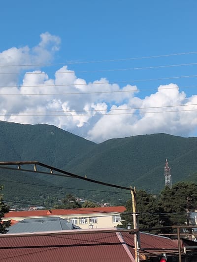 The image shows a view of a mountainside, a blue sky with fluffy white clouds, and some buildings. A communication tower is visible on the mountainside. There are power lines running across the foreground of the image. The buildings appear to be residential or commercial. The scene is outdoors and suggests a sunny day. The time of day is likely daytime due to the bright sunlight. The location cues, such as the architecture of the buildings and the lush greenery on the mountainside, along with the given location context of Şəki, Azerbaijan, contribute to the setting. No people are visible in the image, and no specific activity or event is taking place. There is no visible text in the image.