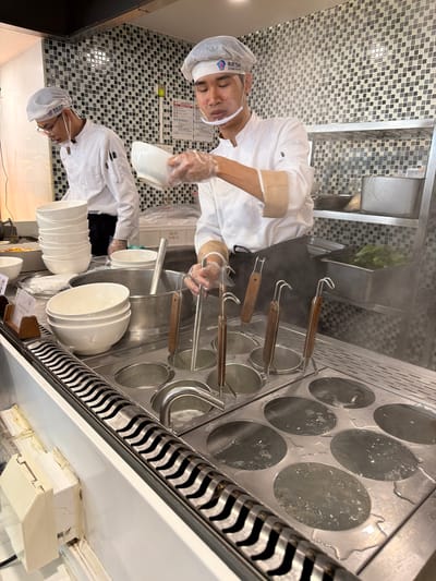 The image depicts an indoor scene within a commercial kitchen, likely located in Baiyun District, China. A chef, dressed in a white chef jacket, black apron, white hairnet, clear face shield, and plastic gloves, is actively engaged in food preparation. The chef is holding a white ceramic bowl, appearing to be in the process of serving or preparing a dish, likely noodles, given the surrounding equipment.
The central part of the scene features a large stainless steel noodle cooker or boiler with multiple circular openings. Several of these openings contain boiling water, from which steam is visibly rising. Other openings hold metal noodle strainers with wooden handles, indicating a noodle-making operation. To the right of the cooker, there's a distinctive metal grate with a wavy pattern. Stacks of clean white ceramic bowls are arranged on the stainless steel counter, ready for use.
The background wall is tiled with small square tiles in a mosaic pattern of black, grey, and white, typical of a professional kitchen. Stainless steel sinks and surfaces are also visible. A reflection in the lower part of the image shows another chef (or the reflection of the same chef) wearing a similar uniform and glasses, reinforcing the impression of a busy food preparation area.
A significant detail is the visible text on the main chef's white hairnet, which reads "南航食品" (Nán Háng Shí Pǐn). This translates to "China Southern Airlines Food," suggesting the establishment is a catering service or restaurant associated with China Southern Airlines. The overall scene conveys a focused and organized environment for food preparation.