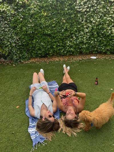 Two young women are lying on a blue and white striped towel on artificial grass. They are both wearing summer clothing and smiling at the camera. The woman on the left is wearing a white and blue striped jumpsuit and has her long brown hair spread out around her. The woman on the right is wearing a red and white floral crop top and black shorts and has her long brown hair spread out around her. They both have their eyes closed and are looking at the camera. A small brown dog is sitting behind them, looking at the camera. There is a bottle of beer on the grass. The location is a grassy area in front of a wall covered in green vines and white flowers. This grassy area is most likely a private outdoor space, such as a patio or backyard. The coordinates (latitude: 40.469, longitude: -3.857) are located in the city of Majadahonda, Spain. There is a wall covered in green vines and white flowers to the left of the image. The wall is slightly out of focus and it appears to be made of brick. The women are the primary subjects of the image. The scene is bright and sunny, and the mood is light and cheerful. The women are lying on their backs, with their legs extended in front of them. The dog is sitting on the grass, looking at the camera. The grass is a bright green, and the overall scene is one of relaxation and enjoyment.