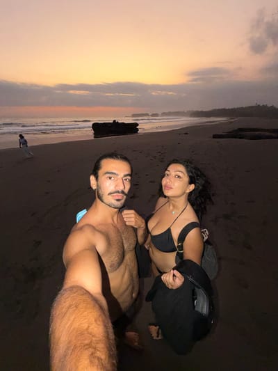 The image depicts a beach scene in Selemadeg Timur, Indonesia, during twilight hours. In the immediate foreground, two individuals are captured in what appears to be a selfie.
The man, positioned on the left, is bare-chested, exhibiting dark hair pulled back, a beard, and a mustache. He wears a small earring in his left ear, and a light blue strap is visible over his left shoulder. His right arm is extended towards the camera, suggesting he is holding the device.
To his right, the woman has dark, curly hair and is wearing a black bikini top with a light-colored necklace featuring a small turquoise pendant. A black strap, likely from a bag, is visible on her left shoulder, and she holds a dark jacket or garment in her right hand. Both individuals are looking directly at the camera.
The background showcases a dark sand beach with gentle waves breaking on the shore. A large, dark rock formation is prominently located in the mid-ground, near the water. Further to the left in the distance, a solitary figure dressed in light-colored clothing stands at the water's edge, facing away from the camera towards the ocean. The sky above displays a gradient of warm orange and yellow hues near the horizon, transitioning into pink and purple tones, with several scattered dark clouds. A dark, vegetated coastline is discernible along the far right horizon. The overall lighting is dim, indicating low natural light conditions, with the foreground subjects appearing distinctly illuminated by a direct light source, likely originating from the camera.