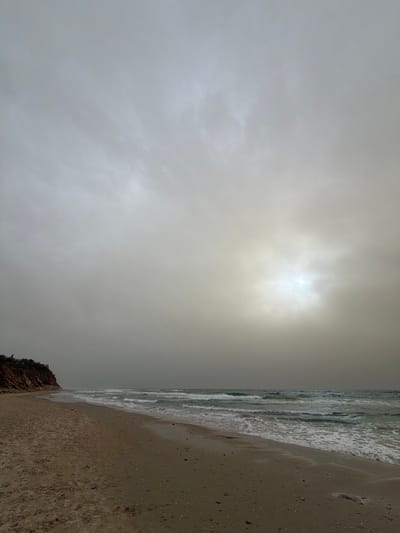The scene depicts a desolate coastal landscape at Beit Yanai, Israel, under an overcast sky. A wide sandy beach occupies the foreground and midground, extending towards the lower left. The sand is light brown-grey and appears damp near the breaking waves. To the left, a prominent, light brown sandstone cliff, topped with sparse green vegetation, runs along the shoreline. The Mediterranean Sea stretches across the middle and right sections of the image, characterized by moderate waves with white foam crests breaking onto the beach. The water displays muted green and blue tones. The sky is entirely covered by a thick layer of grey-white clouds, with a bright, diffuse light source, presumably the sun, partially visible through the cloud cover in the upper right quadrant, casting a pale glow. No individuals, animals, or man-made objects are present in the observable environment.