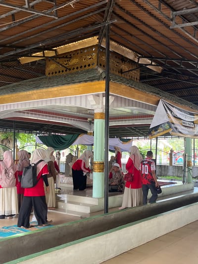 This image captures an outdoor scene at what appears to be a religious site, possibly a tomb or mausoleum, in Beuringen, Indonesia. The foreground features a concrete structure with a green border, possibly a trough or a low wall. Several people are present, mostly women wearing hijabs, dressed in modest clothing, with some opting for long skirts and others for loose-fitting pants. One woman in the center wears a red top with white accents and black pants, carrying a black backpack. Another woman to her right wears a similar red top with white sleeves and a beige hijab. A man in a red and white jersey with the number "10" on the back is also visible.
Behind the people is a raised, open-air structure with a multi-tiered roof supported by columns, some of which are ornately decorated. The roof appears to be made of corrugated material, with elements of gold and intricate designs visible. Fabric drapes, one dark green and one white, are hung within the structure. A banner with Arabic script is also displayed, suggesting a religious context. The background shows greenery and trees, indicating an outdoor environment. The overall atmosphere suggests a place of worship or pilgrimage, with individuals engaged in respectful observation or prayer.