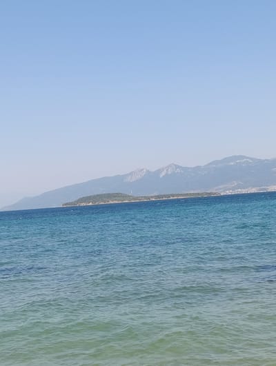 This media file captures a serene seascape under a clear, light blue sky. The foreground is dominated by the tranquil, rippling waters of the sea, which transition from a deep blue in the distance to a lighter, almost aquamarine hue closer to the viewer, suggesting shallowness. The gentle waves create a textured surface on the water.

In the mid-ground, a long, narrow strip of land extends across the horizon. This landmass is densely covered with lush green vegetation, appearing as a verdant island or peninsula. Beyond this green band, a range of mountains rises, their peaks appearing rugged and partially obscured by a light haze, indicating distance and perhaps a warm, sunny day. The mountains exhibit shades of grey and brown, with hints of rocky outcrops visible on their slopes. On the far right of the horizon, a small cluster of buildings, possibly a town or village, can be seen nestled at the base of the mountains.

The scene conveys a sense of peace and natural beauty. The clear sky suggests fair weather and likely daytime, possibly late morning or early afternoon given the brightness. The overall impression is one of a calm, picturesque coastal environment. There is no visible text in the image.